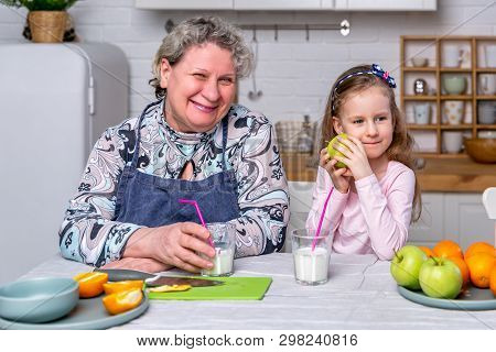 Happy Little Girl And Her Grandmother Have Breakfast Together In A White Kitchen. They Are Having Fu