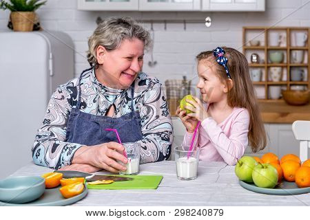 Happy Little Girl And Her Grandmother Have Breakfast Together In A White Kitchen. They Are Having Fu