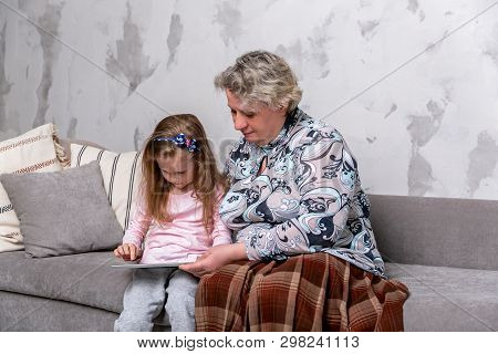 Grandmother And Her Little Granddaughter Are Watching Movies Together And Playing On The Device Whil