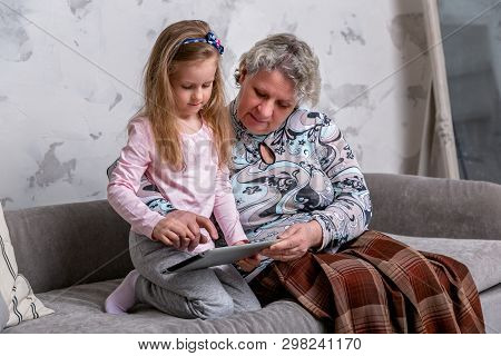 Grandmother And Her Little Granddaughter Are Watching Movies Together And Playing On The Device Whil