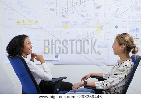Businesswomen Relaxing In Chairs In Front Of Bug Wall Covered With Various Charts, Digrams And Repor
