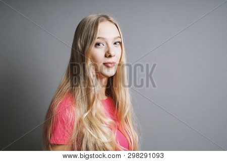Confident Contented Young Teenage Woman With Long Blond Hair And Pink T-shirt Against Gray Backgroun