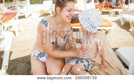 Young Caring Mother With Little Oddler Boy Sitting On Sunbed On Beachand Using Sunscreen Lotion Befo
