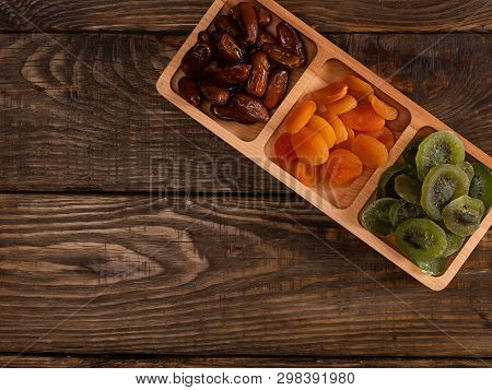 Dates, Dried Apricots And Kiwis In A Compartmental Dish On A Dark Wooden Table.