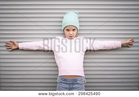 A Serious Girl In A Pink Sweater And Blue Hat Stands Against A Striped Wall And Spreads Her Arms Wid