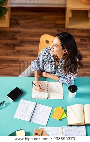 Smiling latin girl looking away while studying. Portrait of pensive college student looking away while studying alone at library. Young thinking woman taking notes at university.