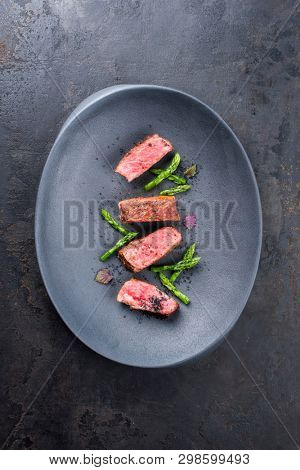 Barbecue dry aged wagyu fillet steak with blanched green asparagus tips and herbs as top view on a modern design plate with copy space