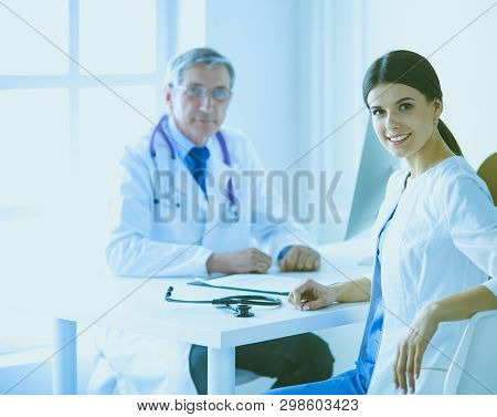 A Doctor Smiling At The Camera With Her Male Colleage In The Back Of The Consulting Room In Hospital