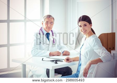 A Doctor Smiling At The Camera With Her Male Colleage In The Back Of The Consulting Room In Hospital