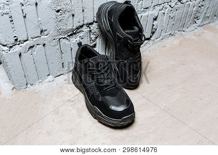 Pair Of Black Sneakers With Thick Soles On The Cement Floor Against The Background Of White Wall