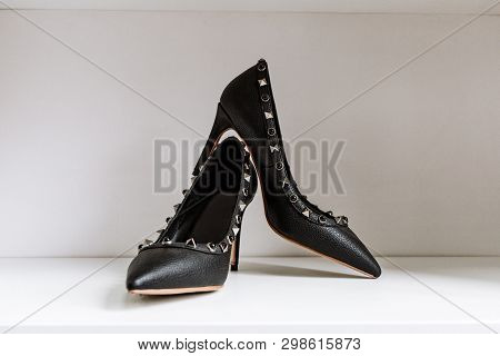 Pair Of Black High-heeled Shoes With Pointed Toes, Decorated With Metal Inserts Against A Shelf In T