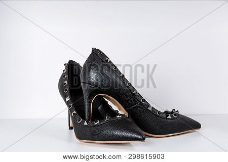 Pair Of Black High-heeled Shoes With Pointed Toes, Decorated With Metal Inserts Against A Shelf In T