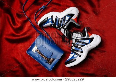 White Sneakers With Black And Blue Inserts On A Thick Figured Sole And A Blue Bag With A Gold Chain