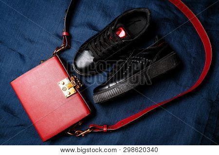 Black Sneakers With Red Tongues And A Red Bag With A Golden Lock On A Blue Woven Background