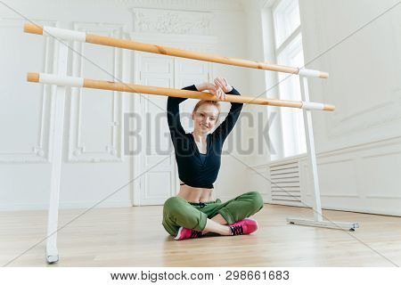 Pleased Young Female Dancer Sits In Lotus Pose, Keeps Hands On Ballet Barre, Has Rest After Choreogr