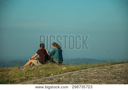 Guarda, Portugal - July 16, 2018. Couple Of Young People Sitting On Lawn With Hilly Landscape On Sun