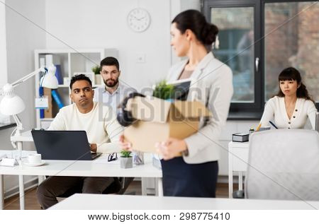 business, firing and job loss concept - sad fired female employee with box of personal stuff leaving office looking at her colleagues