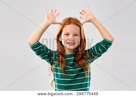 childhood, gesture and people concept - smiling red haired girl in green striped shirt having fun and making big ears over grey background