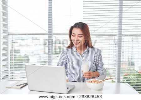 Beautiful Young Woman Sitting At The Table In Home Office, Holding Glass Of Milk With Cereal, Using 