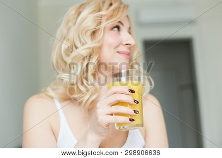 Attractive Blonde Woman Sits On The Edge Of The Bed At Home And Holds A Stocker With Orange Juice In