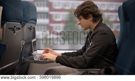 Young Guy With Laptop Sitting In Train, Student In Black Jacket Studying While Traveling. Young Man 