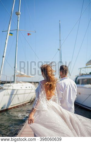 The Newlyweds Walk Near The Yacht Club. The Husbend Leads The Wife By The Hand On The Pier Between T