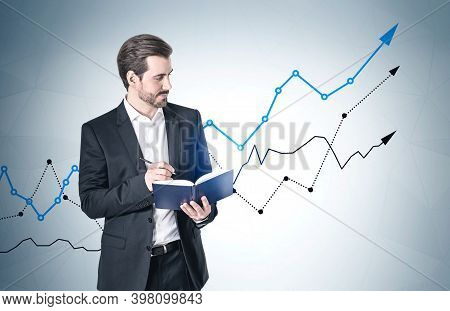Portrait Of Confident Young Businessman Standing With Book Near Gray Wall With Growing Graphs. Conce