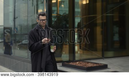 Man In Black Coat And Eyeglasses Looking At The Phone Walking Along The Street With Coffee, Slow Mot