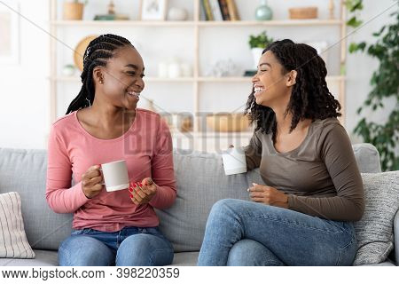 Happy Black Girlfriends Enjoying Weekend, Drinking Coffee And Talking, Sitting On Couch At Home, Sha
