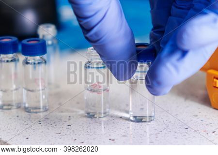 Close Up Scientist Hand In Rubber Gloves Takes A Glass Vial With A Blue Cap For Hplc Analysis. Devel