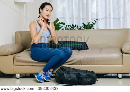 Pretty Young Asian Woman In Sports Clothes Sitting On Sofa In Living Room And Putting On Headphes To