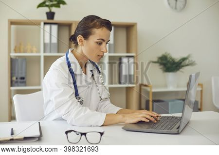Erious Female Practitioner Typing On Laptop Keyword Sitting At Desk In Office Of Modern Clinic