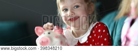 Portrait Of Smiling Charming Child Holding Piggy Bank Sitting On Sofa Indoors. Lovely Blonde Kid Sav