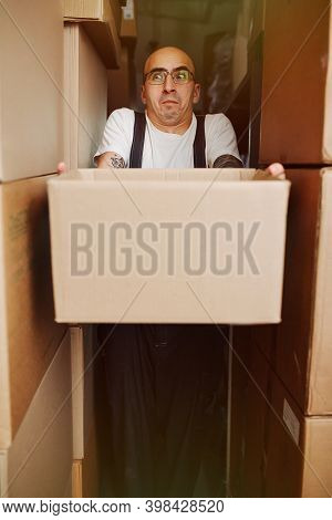Funny Warehouse Worker Carrying Box, Squeezes Between Tall Raws Of Stacked Boxes
