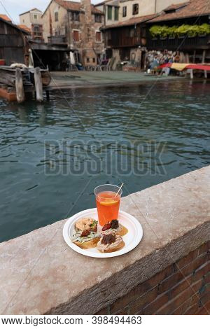 Traditional Venetian Food And Drink Cicchetti And Aperol Spritz With Turquoise Green Water Of Canal.