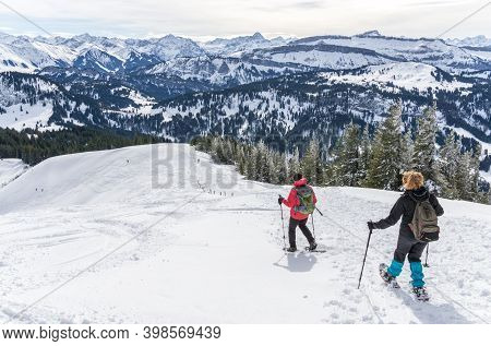 Senior Couple Is Snowshoe Hiking In Alpine Snow Winter Mountains. Allgau, Bavaria, Germany.