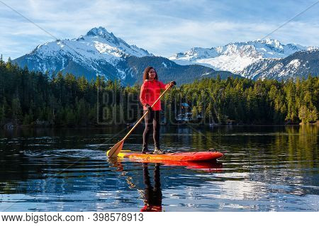 Adventurous Girl Paddle Boarding On Levette Lake With Famous Tantalus Mountain Range In The Backgrou