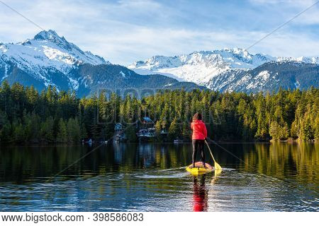 Adventurous Girl Paddle Boarding On Levette Lake With Famous Tantalus Mountain Range In The Backgrou