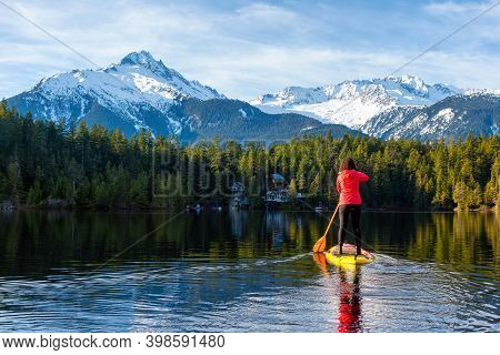 Adventurous Girl Paddle Boarding On Levette Lake With Famous Tantalus Mountain Range In The Backgrou