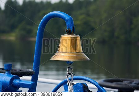 Brass Bell On The Foredeck Of A Ship, Close-up Against The Background Of A Blurred Wooded Coast