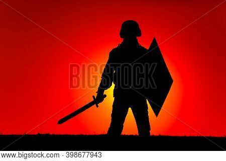 Silhouette Of Modern Soldier In Combat Helmet, Armed Sword And Shield, Standing On Background Of Sun