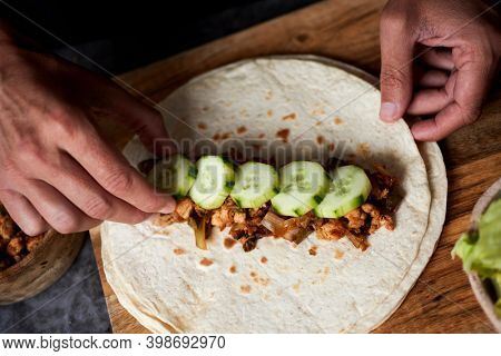 high angle view of a young man preparing a durum or a burrito, with chicken meat cooked with different vegetables such as onion or red and green pepper, and fresh lettuce and raw cucumber