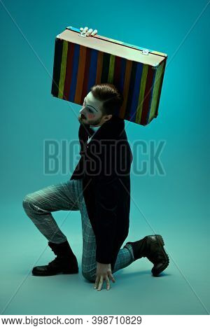 Art and fashion. Portrait of a young man with makeup in the style of the 20s in an elegant suit standing with his old suitcase. Male mime artist. Studio portrait.