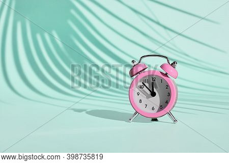 Pink Alarm Clock On Light Blue, Turquoise Pastel Background With A Shadow Of Palm Leaf. Summertime, 
