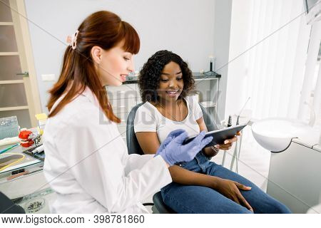 Portrait Of Woman Dentist Holding Tablet, While Demonstrating Smth To Her Young Female African Patie