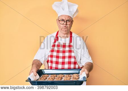 Senior grey-haired man wearing baker uniform holding homemade cookies puffing cheeks with funny face. mouth inflated with air, catching air. 