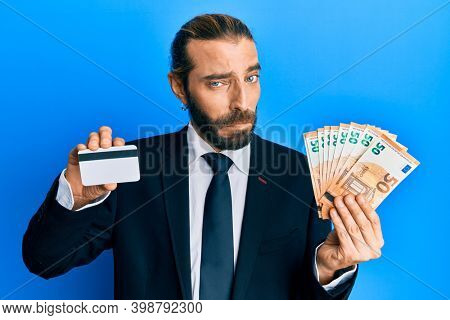 Attractive business man with long hair and beard holding credit card and euros skeptic and nervous, frowning upset because of problem. negative person. 