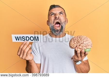 Middle age handsome man holding wisdom word on paper and brain angry and mad screaming frustrated and furious, shouting with anger looking up. 