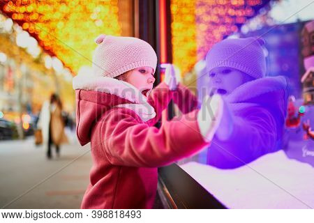 Toddler Girl Looking At Window Glass Of Large Department Store Decorated For Christmas. Child Enjoyi
