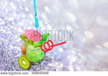 A Glass Of Green Drink With Ice, Decorated With A Cocktail Umbrella And Straws On A Gray Abstract Ba